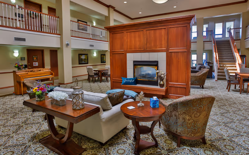 A spacious and well-lit senior living facility common area featuring a central wooden fireplace with a 'Grateful' pillow on the hearth. Surrounding the fireplace are comfortable seating options including a beige sofa with cushions, patterned armchairs, and wooden tables adorned with decorative items and flowers. The room has a patterned carpet, high ceilings, and a second-floor balcony overlooking the area. Stairs and additional seating are visible in the background.