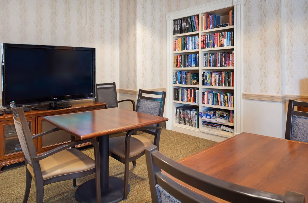 A cozy room with two wooden tables surrounded by chairs, a large flat-screen TV on a wooden cabinet, and a built-in bookshelf filled with books and board games against a wallpapered wall.