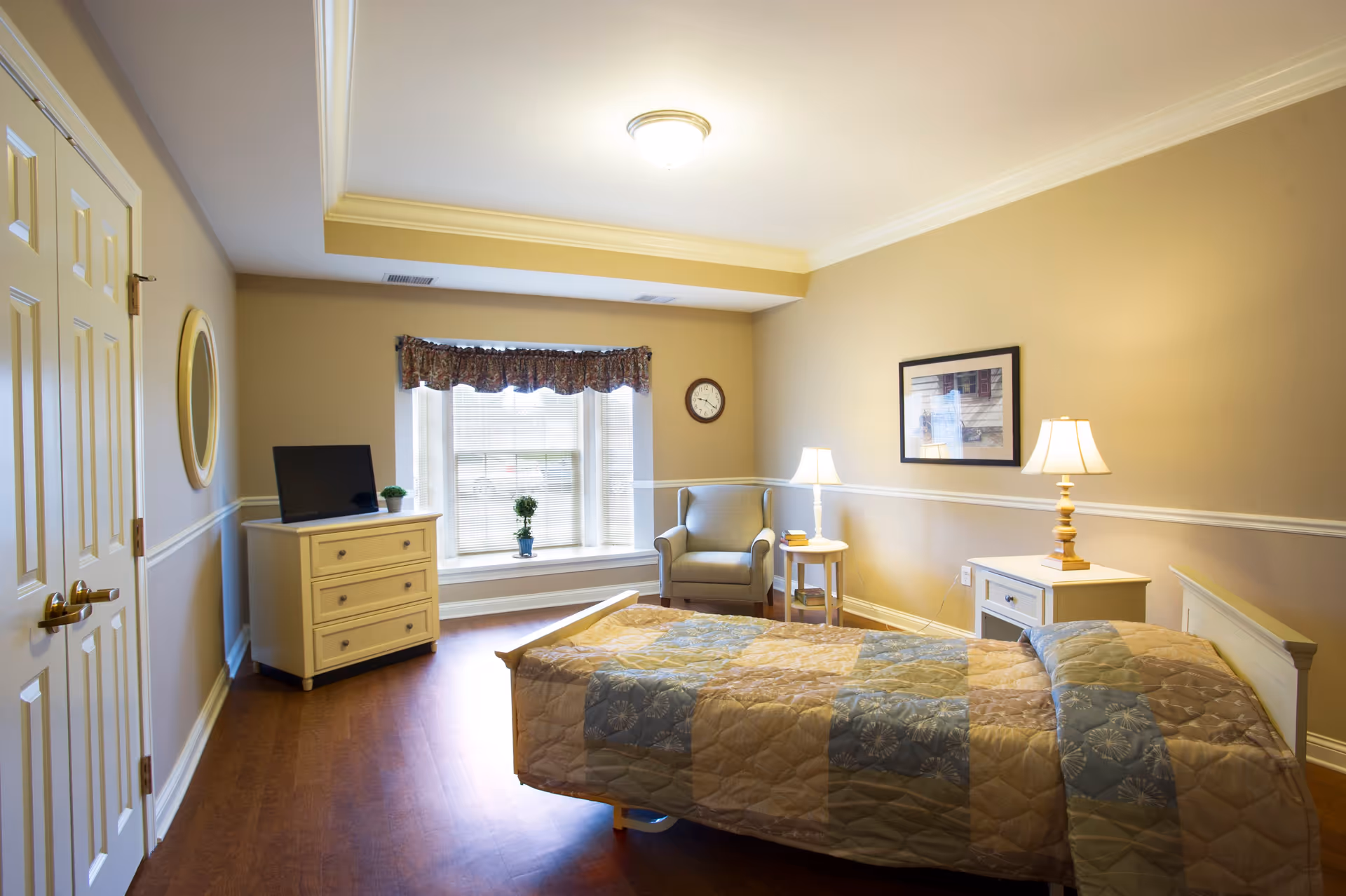 A well-lit bedroom with a single bed covered in a patchwork quilt, a small dresser with a TV and a plant on top, a comfortable armchair next to a window with blinds and a floral valance, a side table with a lamp and books, and a framed picture on the wall.