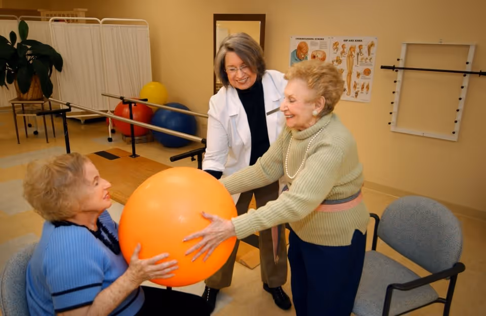 Two elderly women participating in a physical therapy exercise with a large orange exercise ball, assisted by a female therapist in a clinical setting with exercise equipment and anatomical posters in the background.