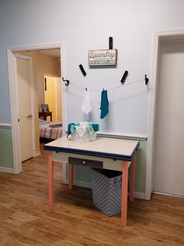Interior hallway area with a small table holding a laundry basket beneath a decorative 'Laundry' sign and clothespins with hanging towels.