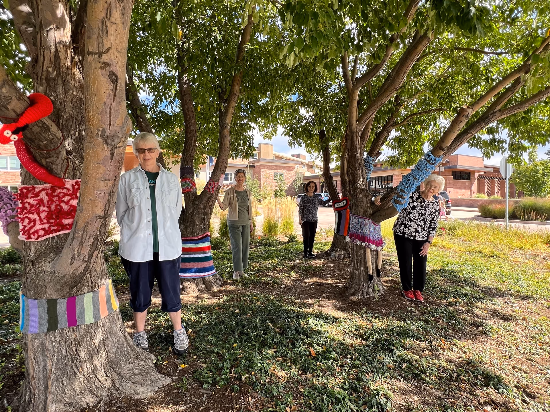 Four elderly women standing outdoors among trees that are decorated with colorful knitted or crocheted coverings on their trunks and branches. The women are smiling and posing in a garden area with a building visible in the background under a partly cloudy sky.