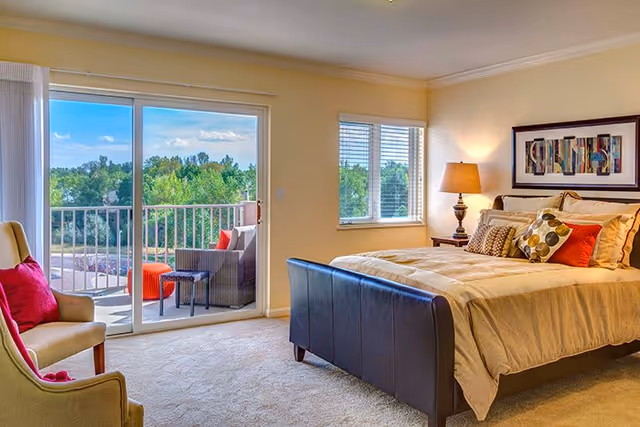 A bright bedroom with a large bed featuring multiple pillows, a bedside table with a lamp, an armchair with a red cushion, and a sliding glass door leading to a balcony with outdoor seating and a view of trees.