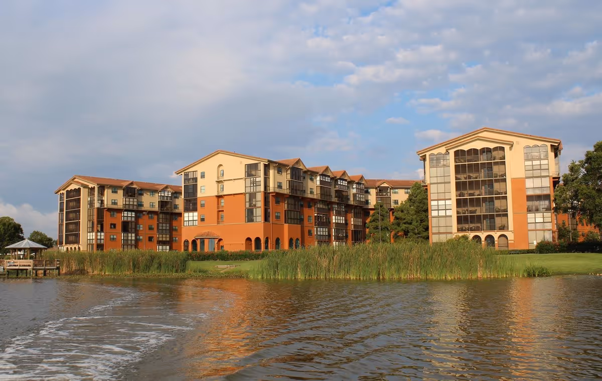 View of a multi-story senior living facility building named Lake Seminole Square, situated beside a body of water with tall grass along the shoreline under a partly cloudy sky.