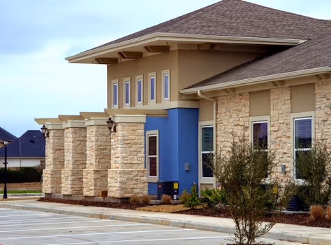 Exterior view of a senior living facility building with stone and beige walls, blue accent panels, multiple windows, and a sloped roof. There are small trees and shrubs planted along the building, and an empty parking lot in the foreground.