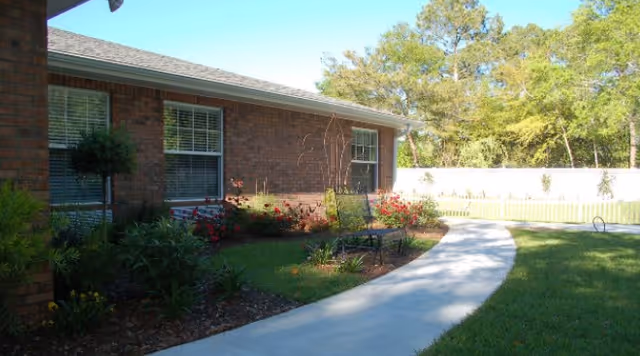 A curved concrete walkway leading alongside a brick building with windows, bordered by green grass, bushes, and flowering plants. Trees and a white fence are visible in the background under a clear blue sky.