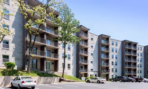 Exterior view of a multi-story apartment building with balconies, trees, and a parking lot with several parked cars under a clear blue sky.
