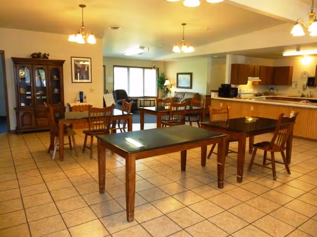 A spacious room with tiled flooring featuring several wooden tables and chairs arranged for dining or activities. In the background, there is a kitchen area with wooden cabinets, a stove, and a microwave. A living area with a sofa and armchair is visible near a window with blinds. The room is well-lit with ceiling lights and has a china cabinet against one wall.