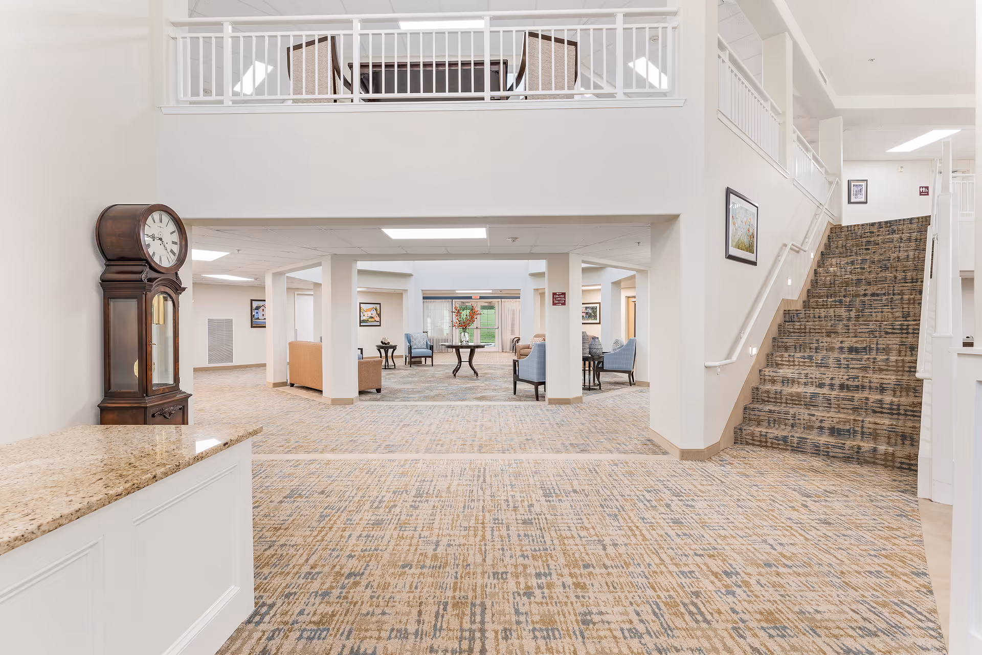 Bright open lobby of a senior living facility with seating areas, a grandfather clock, and a carpeted staircase.