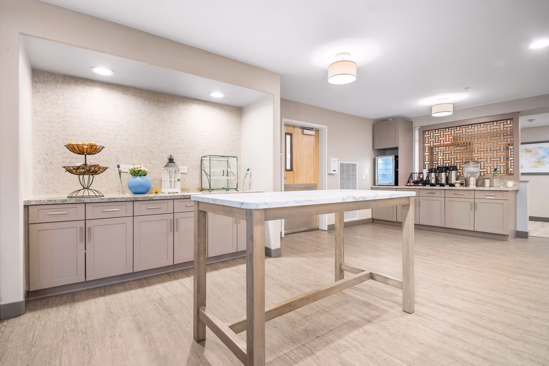 A bright and modern common area with light wood flooring, beige walls, and recessed lighting. There is a tall table with a marble top in the center, cabinets along the walls with decorative items including a tiered fruit basket, a blue vase with flowers, a lantern, and a glass display case. On the right side, there is a coffee and beverage station with multiple coffee dispensers and cups, set against a decorative wooden lattice panel.
