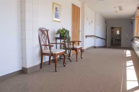 Quiet interior hallway with wooden chairs, a plant, and framed art along a sunlit corridor.