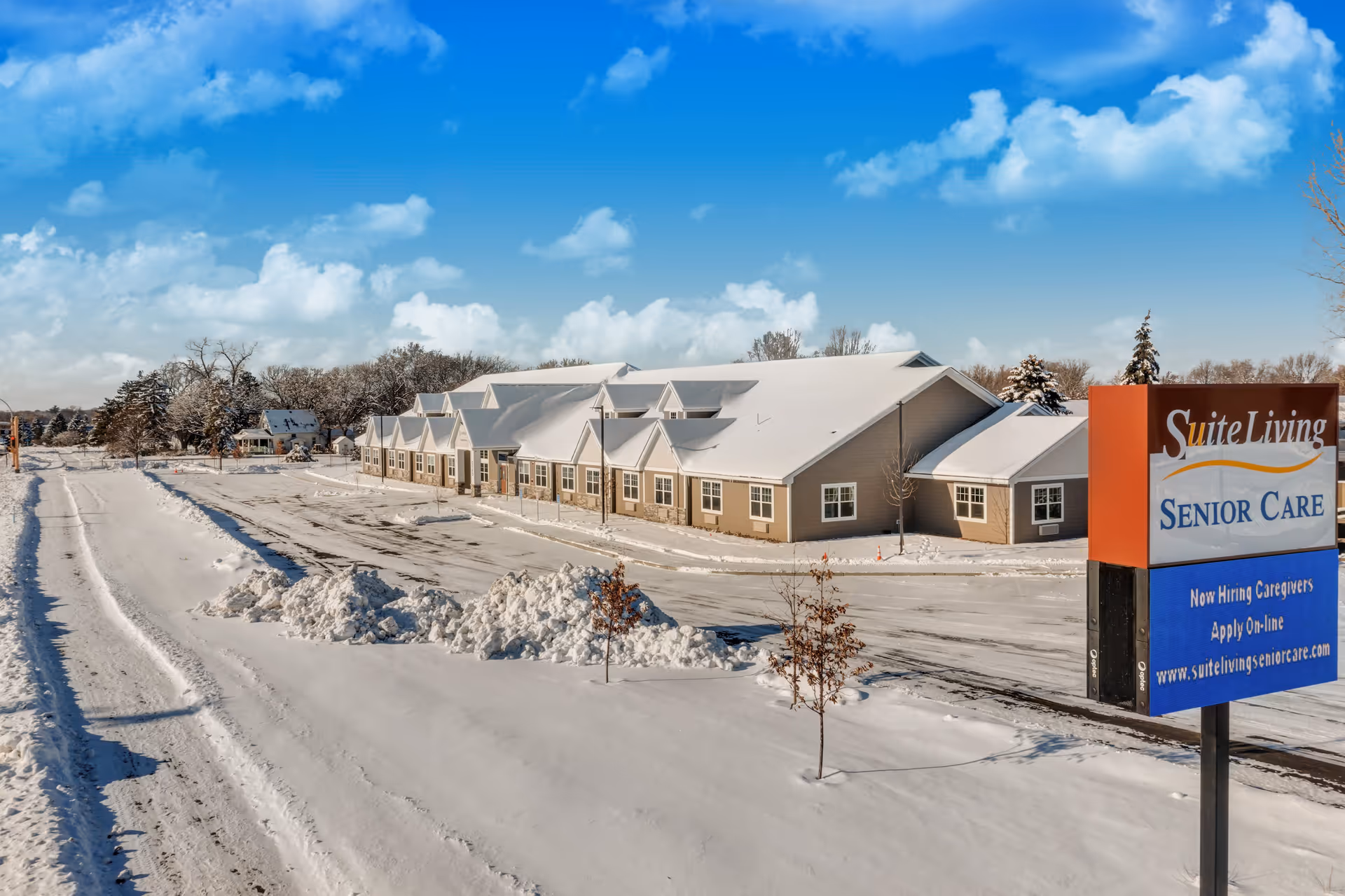 Exterior view of Suite Living of Spring Lake Park senior care facility covered in snow on a clear winter day with a blue sky and scattered clouds. A large sign in the foreground advertises hiring caregivers and provides the website www.suitelivingseniorcare.com.
