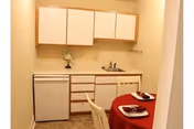 Small kitchenette area with white cabinets, a sink, a mini refrigerator, and a countertop. In the foreground, there is a round table covered with a red tablecloth, set with two plates and utensils.