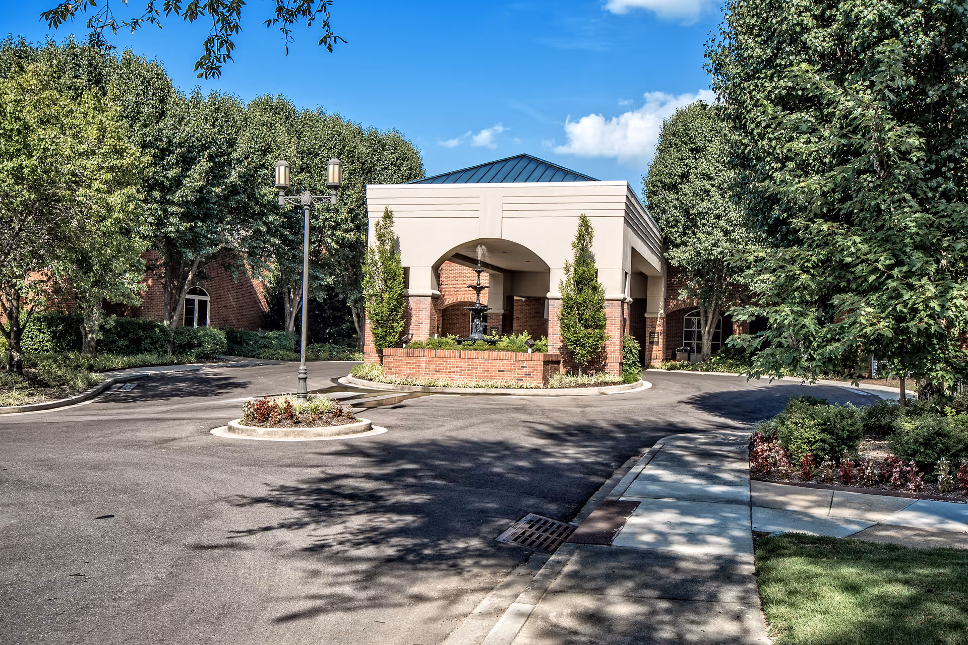 Entrance area of a senior living facility with a covered drop-off zone, surrounded by trees and landscaped greenery under a clear blue sky.