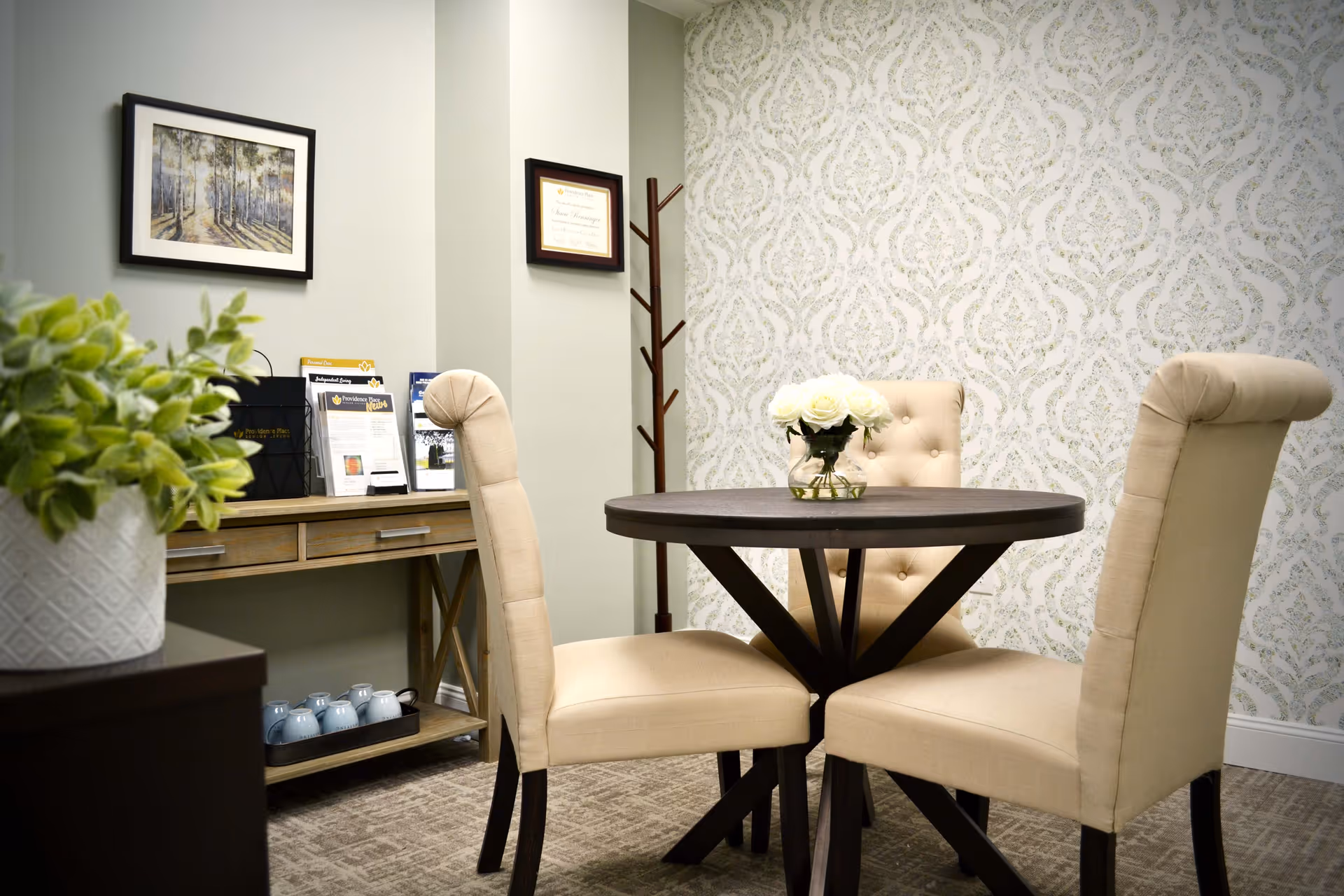 A small round wooden table with a vase of white flowers on top, surrounded by three beige upholstered chairs with button tufting. The room has patterned wallpaper on one wall and a light green painted wall with framed artwork and certificates. A wooden console table with brochures and a plant is visible in the background.