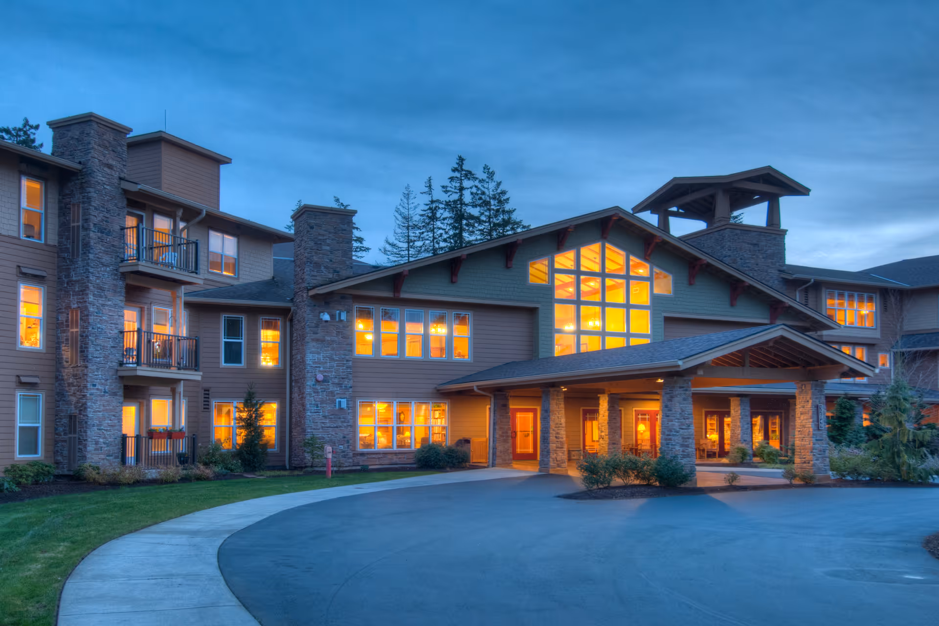 Exterior view of Wesley Lea Hill senior living facility at dusk with warm lights glowing from the windows, a covered entrance supported by stone pillars, and a curved driveway in front.