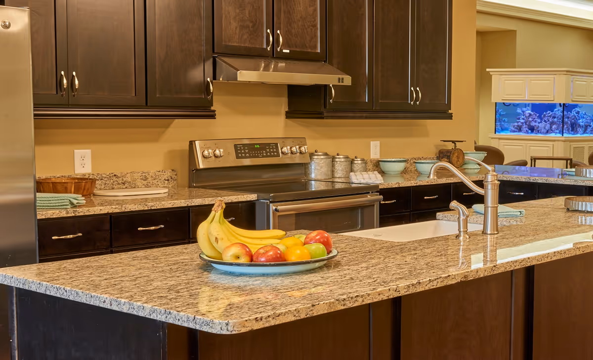 Modern kitchen with a granite island, stainless steel appliances, dark cabinets, and a bowl of fruit on the counter.