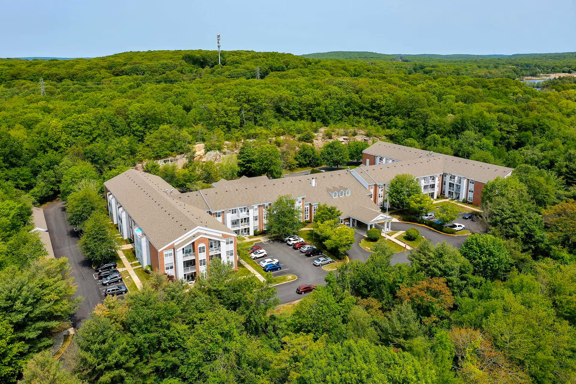 Aerial view of Solstice Senior Living at Groton, showing a large, multi-wing building surrounded by dense green trees and parking lots with several cars. The building has a beige roof and brick exterior with white trim, set against a backdrop of forested hills under a clear blue sky.