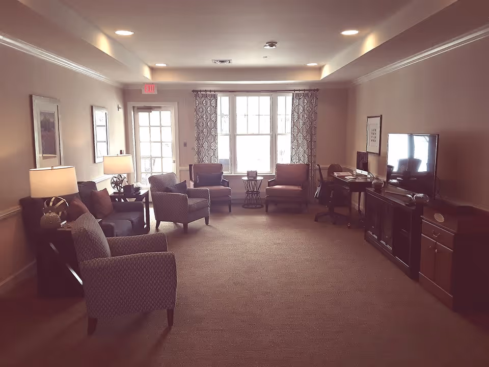 A cozy living room area with multiple armchairs and a sofa arranged around side tables with lamps. There is a large window with patterned curtains letting in natural light. On the right side, there is a TV on a wooden stand and a desk with a computer and office chair. The walls are decorated with framed artwork.