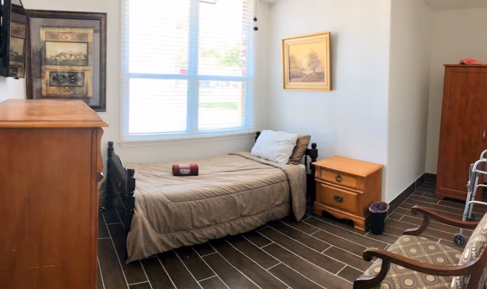 A small bedroom with a single bed covered in a beige comforter and pillows, a wooden nightstand with two drawers, a wooden dresser, a patterned armchair, a walker, a trash bin, and two framed pictures on the walls. A large window with blinds lets in natural light.