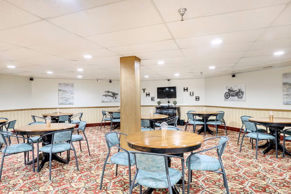 A common area with multiple round wooden tables and teal cushioned chairs arranged on a patterned carpet. The walls are decorated with framed pictures and a TV mounted above a black cabinet with the words 'THE HUB' displayed on the wall behind it. The ceiling has recessed lighting and a wooden pillar stands in the center of the room.