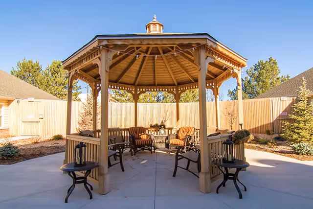 Outdoor wooden gazebo with a peaked roof and open sides, furnished with cushioned chairs and small tables, situated on a concrete patio surrounded by a wooden fence and trees under a clear blue sky.