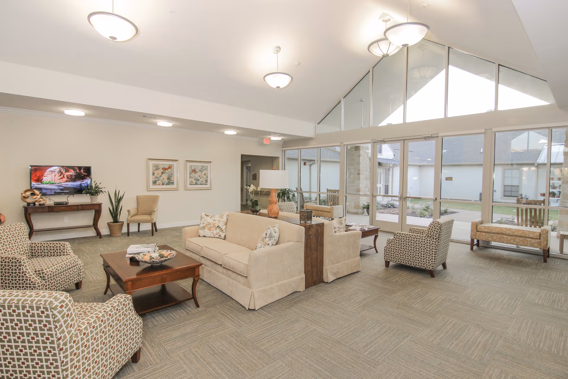A spacious and well-lit living room area with beige sofas, patterned armchairs, a wooden coffee table, and a TV mounted on the wall. Large windows and glass doors provide a view of an outdoor courtyard with benches and greenery. The room has a high ceiling with modern light fixtures and neutral-colored carpeting.