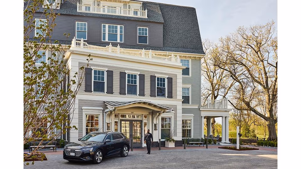 Front exterior view of The Newbury Of Brookline, a multi-story building with gray and white siding, multiple windows, and a covered entrance. A black car is parked in front, and a person is walking near the entrance. Trees and landscaping surround the building.