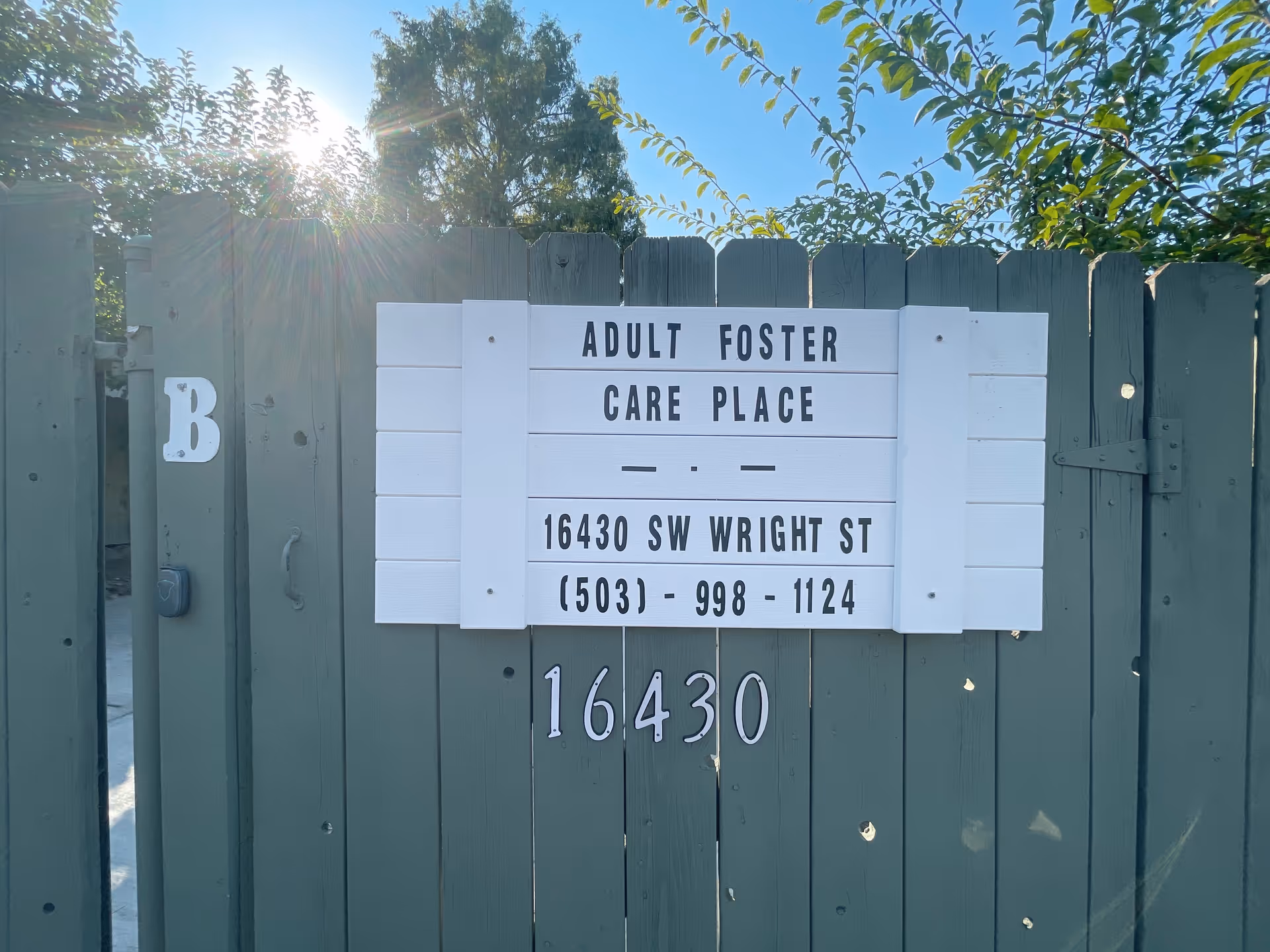 Green wooden fence with a white sign that reads 'Adult Foster Care Place' and shows the address 16430 SW Wright St.