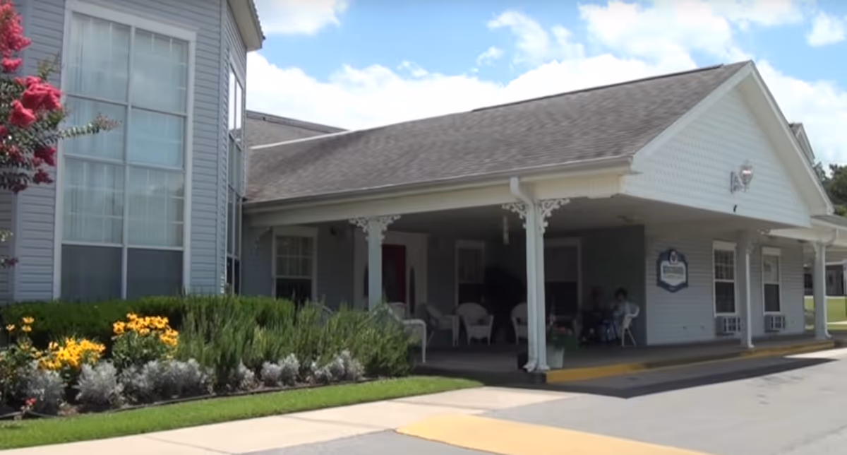 Front covered entrance and porch of a senior living building with chairs, landscaping, and a driveway.