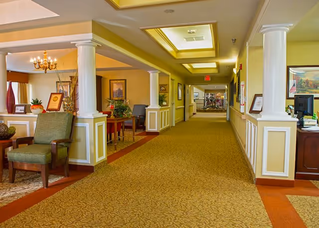 A well-lit hallway in a senior living facility with beige walls, carpeted floor, and white columns. There are chairs and tables with decorative plants and framed pictures along the sides. The ceiling has recessed lighting and a chandelier is visible in a seating area to the left.