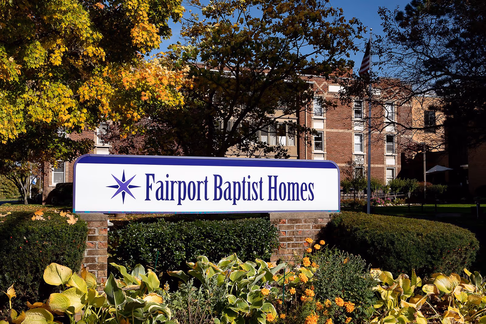 Outdoor view of the Fairport Baptist Homes facility sign surrounded by greenery and flowers, with a multi-story brick building and trees in the background under a clear blue sky.