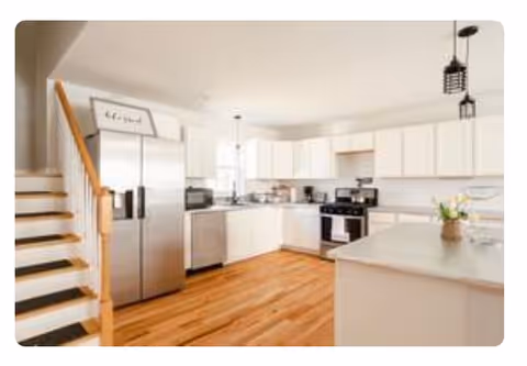 Bright and spacious kitchen with white cabinets, stainless steel refrigerator, oven, microwave, and dishwasher. Wooden floor and staircase with wooden railing on the left side. Kitchen island with a vase of flowers on the right side.