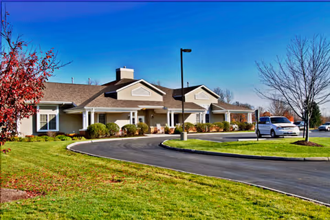 Single-story building with a beige exterior and white trim, surrounded by green grass and a curved driveway. There are a few small trees and bushes around the building, and a white vehicle is parked near the entrance under a clear blue sky.