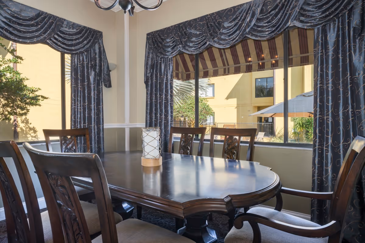 A dining room with a dark wooden table surrounded by six matching chairs. The room has large windows with dark blue curtains featuring a subtle swirl pattern. Outside the windows, there is a view of a yellow building, greenery, and an outdoor umbrella.