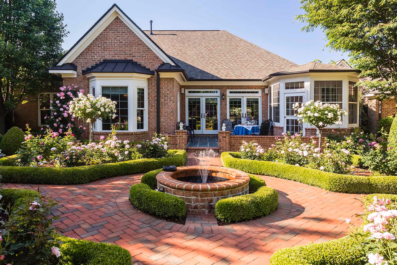 Brick building front with a sunlit courtyard featuring a circular fountain, manicured hedges and rose gardens.
