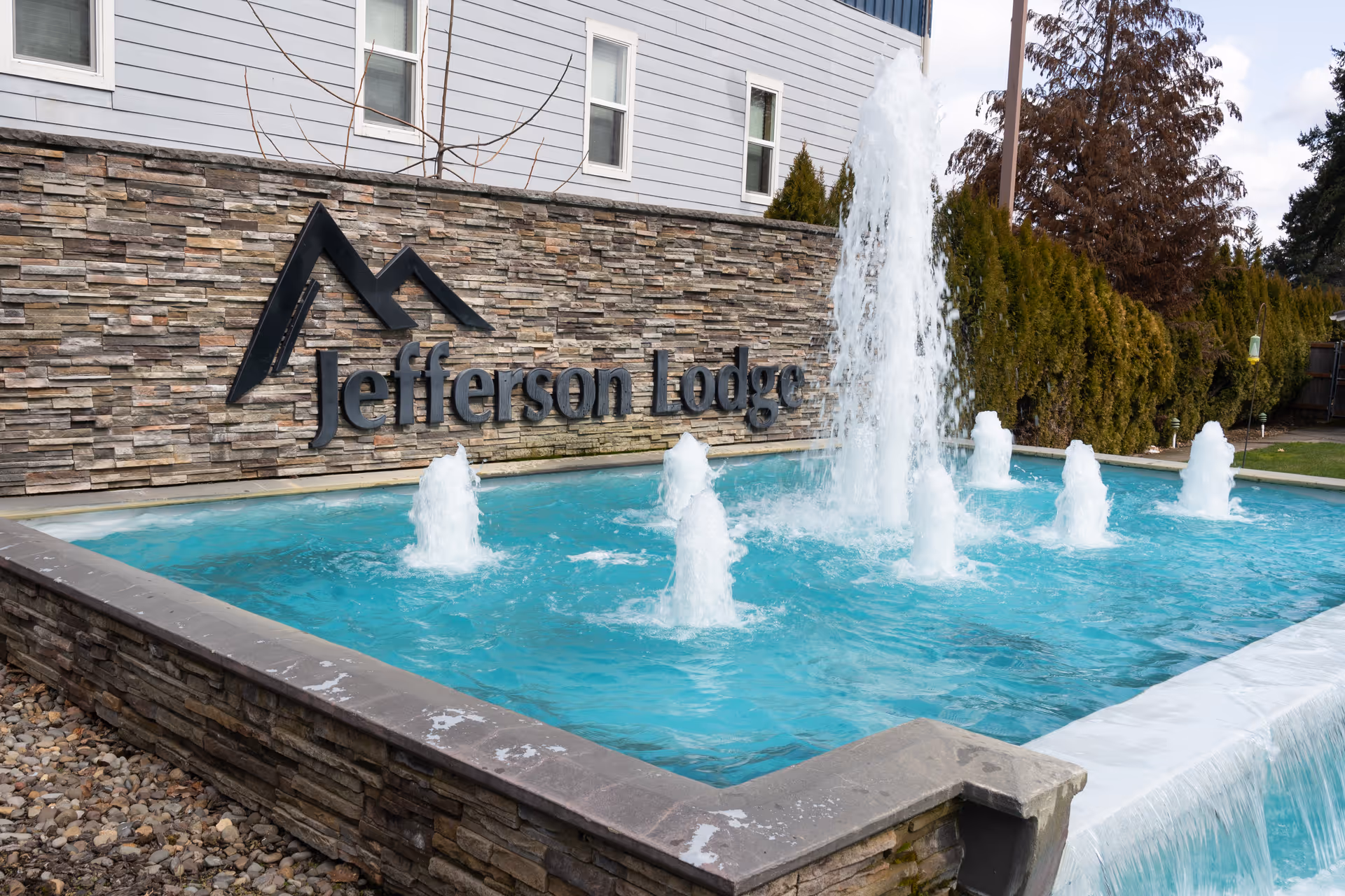 Outdoor water fountain with multiple jets of water in front of a stone wall featuring the sign 'Jefferson Lodge'. The building exterior and some trees are visible in the background.