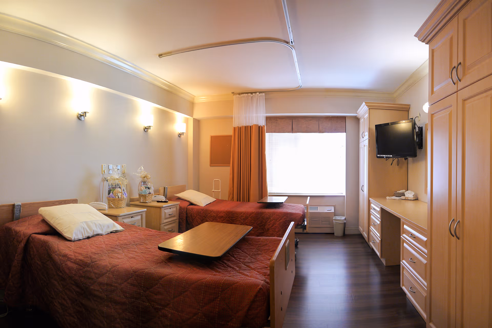 A well-lit senior care facility bedroom with two single beds covered in red quilts, each with a white pillow and an overbed table. There are wall-mounted lights above the beds, a window with blinds and curtains, a mounted flat-screen TV, and wooden cabinetry with drawers and a desk area.