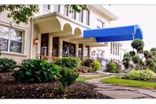 Exterior view of Sterling Health Care & Rehabilitation Center showing a beige building with large windows, a blue awning over the entrance, and well-maintained landscaping including bushes and small trees along a stone pathway.