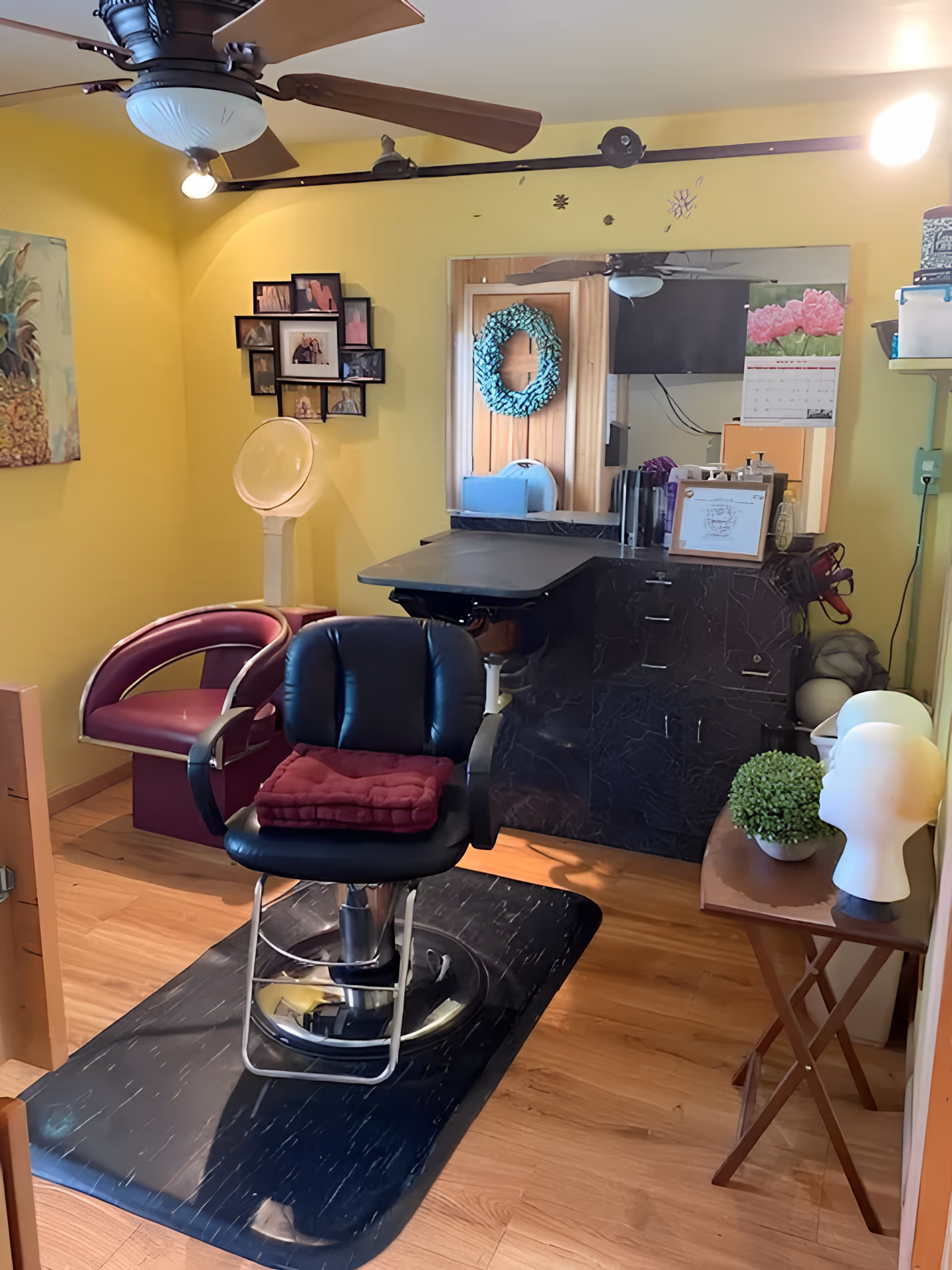 Interior of a small hair salon or barber station with a black salon chair on a black mat, a maroon chair, a hair dryer hood, a large mirror on a yellow wall, a ceiling fan with lights, and a small table with mannequin heads and a potted plant.