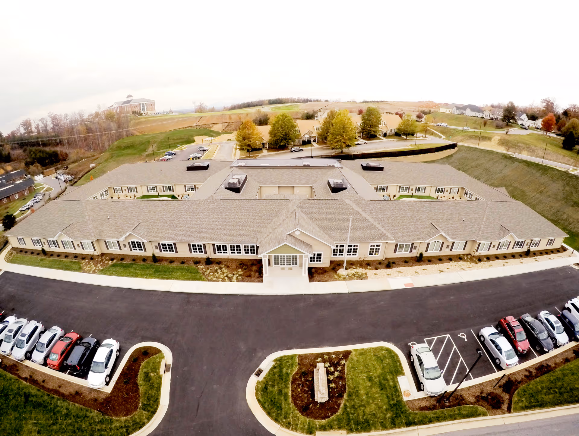 Aerial view of a large single-story senior living facility building with a central entrance, surrounded by parking lots with several cars and landscaped areas. The building is situated in a semi-rural area with open fields and other buildings in the background.