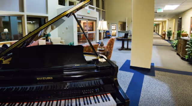 Carpeted lobby/common area with a black Suzuki grand piano in the foreground, seating, lamps, and a hallway.