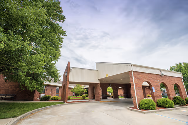 Exterior view of a brick building with a covered entrance and arched openings, surrounded by green bushes and trees under a partly cloudy sky.