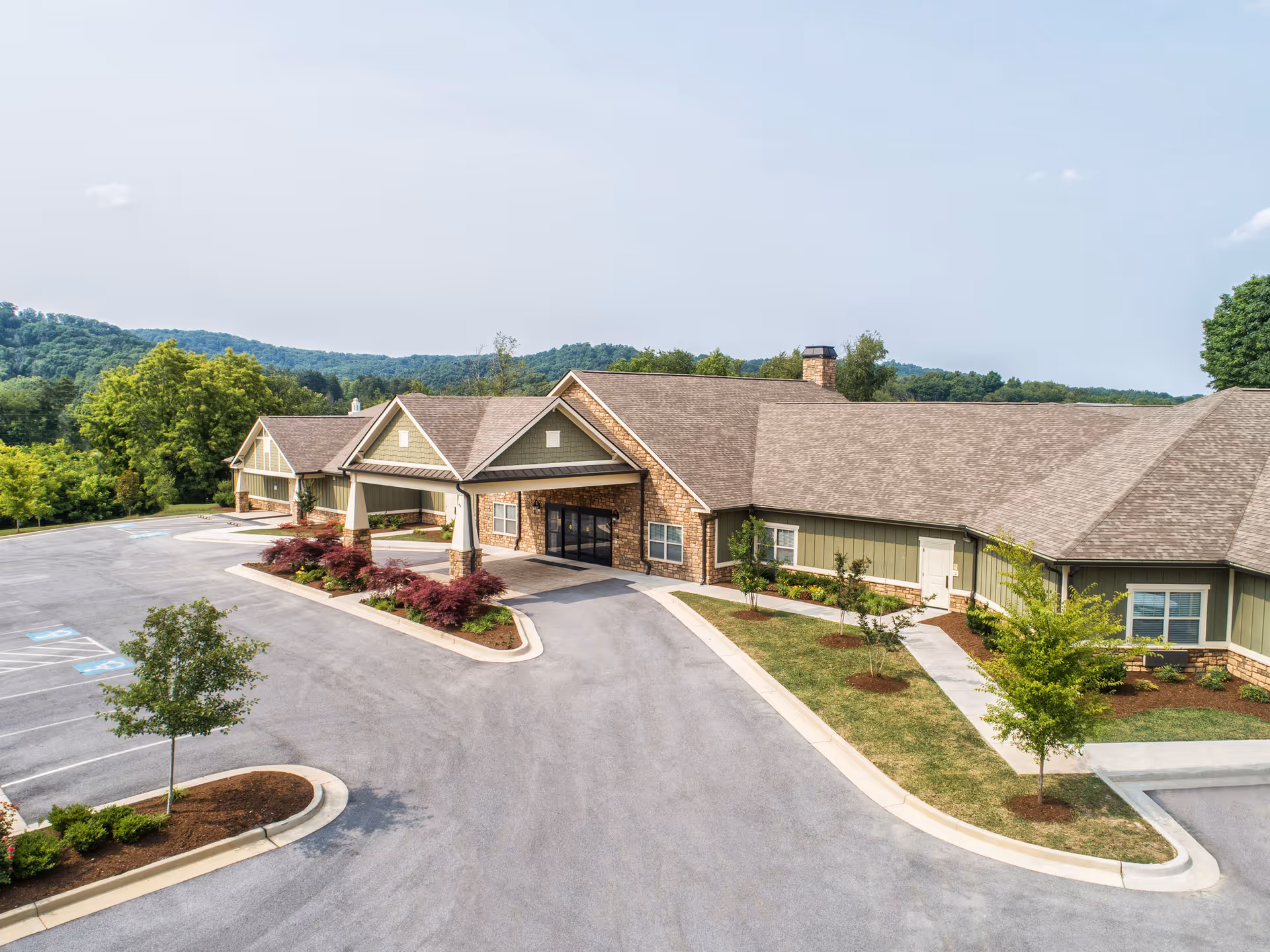 Exterior view of Dominion Senior Living of Bristol, showing a single-story building with a covered entrance, stone and green siding, surrounded by landscaped greenery and a parking lot with handicap spaces.