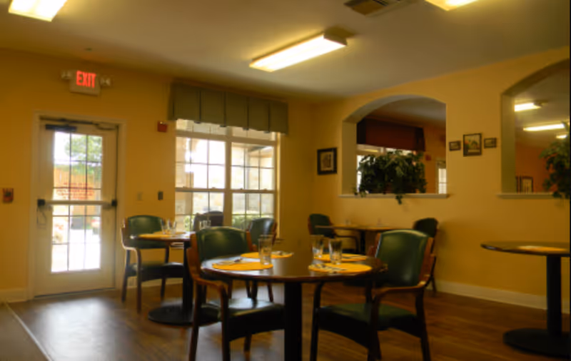 Interior view of a dining room in a senior living facility with round tables set with yellow placemats, glasses, and silverware. Green cushioned chairs surround the tables. There is a glass door with an exit sign above it and large windows letting in natural light. The walls are painted yellow with framed pictures and a large mirror.