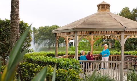 Three elderly women sitting and standing in a wooden gazebo surrounded by lush greenery with a water fountain and trees in the background at a senior living facility.