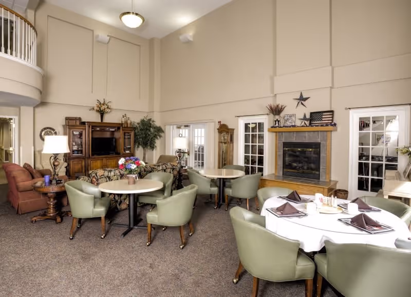 A spacious senior living common area with multiple round tables surrounded by green cushioned chairs. One table is set with a white tablecloth, brown napkins, and cups. The room features a fireplace decorated with stars and an American flag, a wooden grandfather clock, a large wooden entertainment center with a TV, and comfortable seating including a floral sofa and red armchairs. The walls are beige with high ceilings and there are glass doors leading to other rooms.