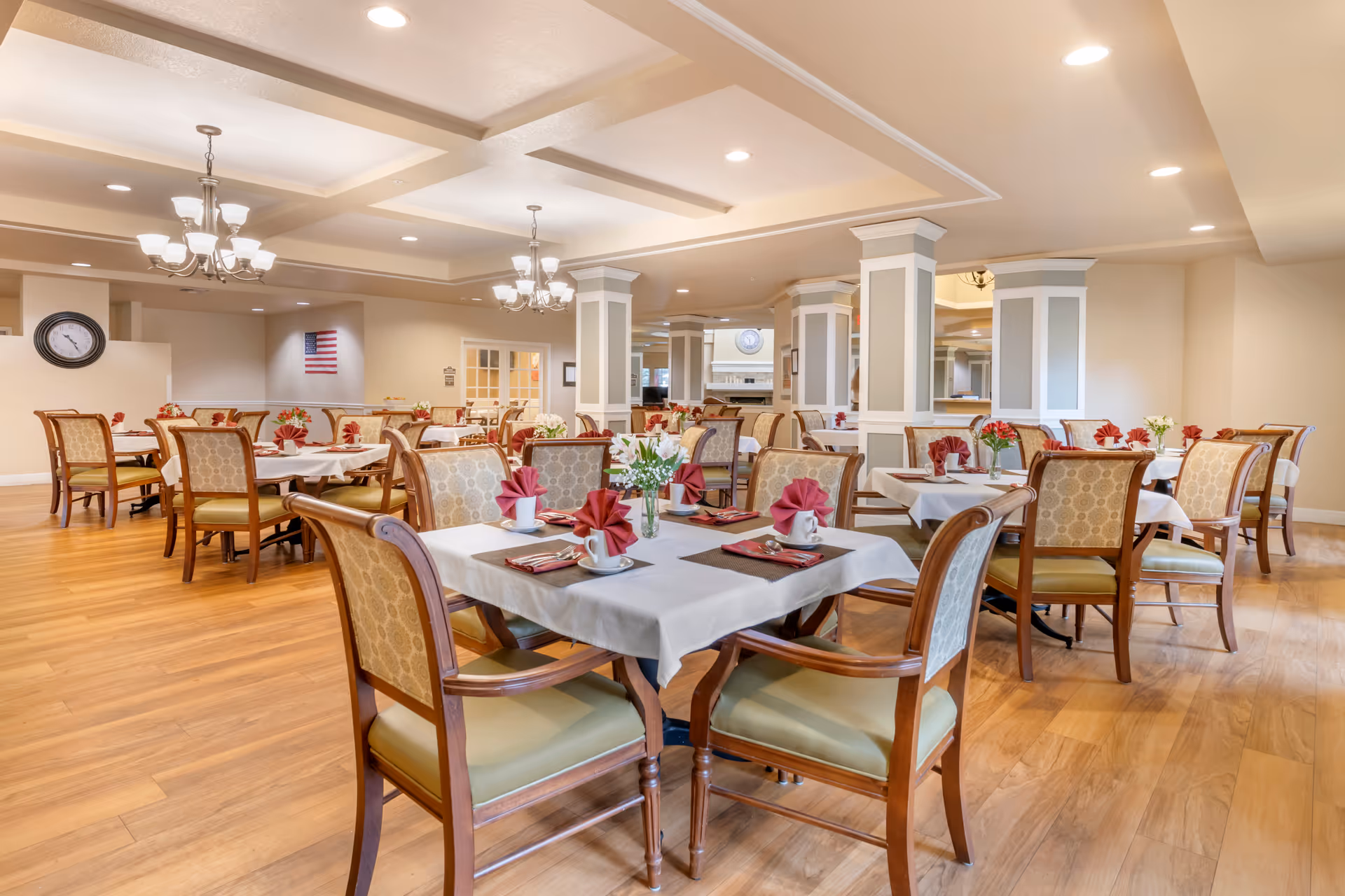 A spacious dining room with multiple tables covered in white tablecloths, each set with folded red napkins, cups, and silverware. The room features wooden chairs with patterned upholstery, hardwood flooring, and soft overhead lighting from chandeliers and recessed lights. There is an American flag on the wall and a clock visible in the background.