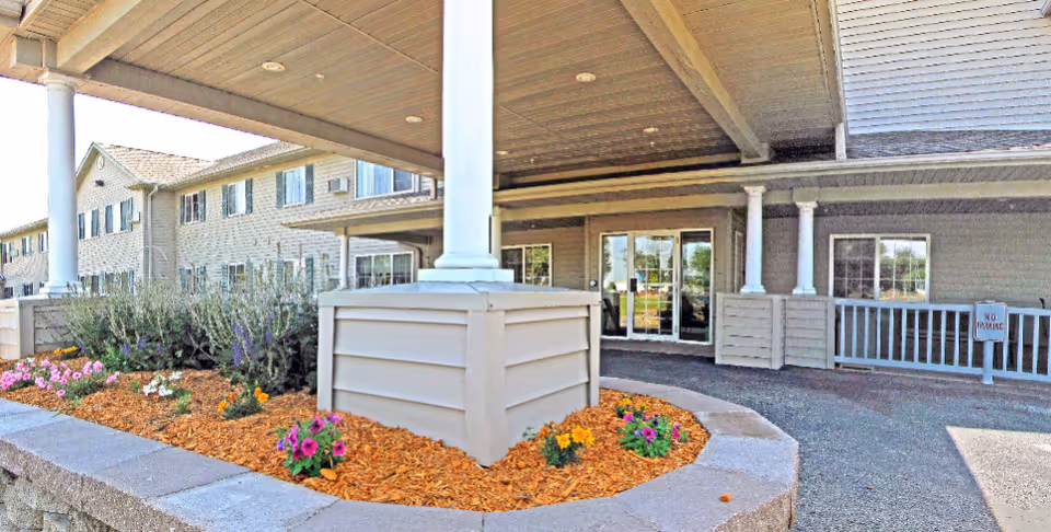 Entrance area of a senior living facility with a covered drop-off zone supported by white columns. There is a landscaped flower bed with mulch and colorful flowers in front of the building. The building has beige siding and multiple windows, with glass double doors at the entrance.
