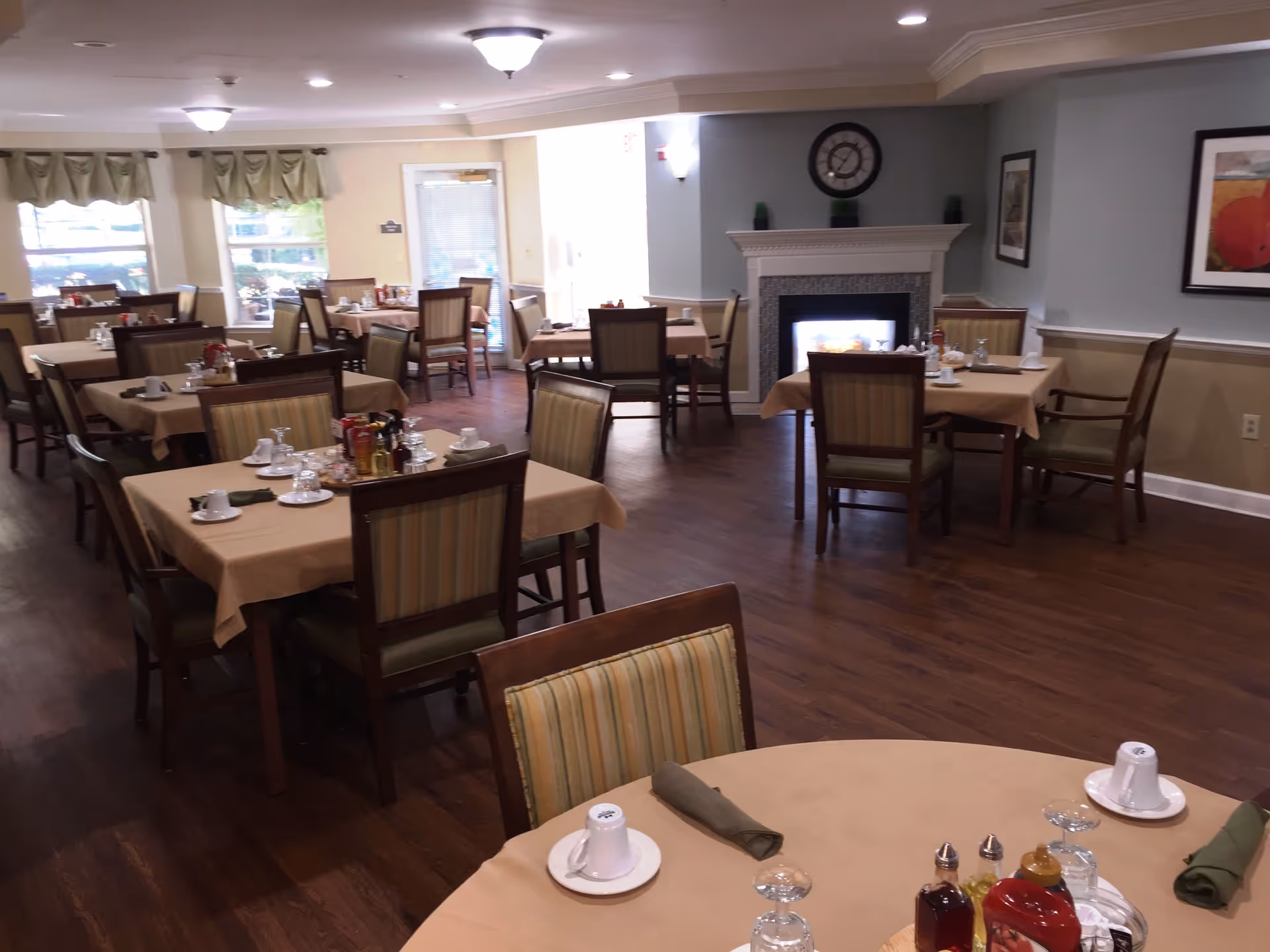 A dining room with multiple tables covered in beige tablecloths, each set with cups, glasses, napkins, and condiments. The room has wooden flooring, a fireplace with a clock above it, framed artwork on the walls, and windows with curtains allowing natural light to enter.